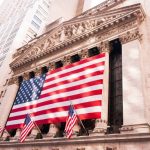 A imagem mostra um prédio de Wall Street dando foco em sua arquitetura. Há uma bandeira grande dos Estados Unidos sobre a fachada e mais três bandeiras pequenas hasteadas em macros.