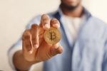 A imagem mostra um homem segurando uma moeda Bitcoin, e ilustra a matéria que fala sobre cuidados para não cair em golpes de Bitcoin.