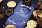 Smartphone, ao lado de moedas,exibe o nome bitcoin na tela