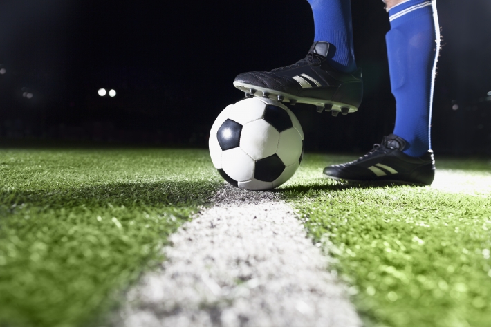 Imagem mostra detalhe de pé sobre bola de futebol, posicionada em cima da linha de cal branca.