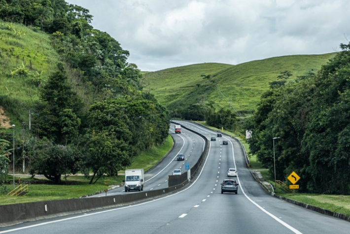 a imagem mostra uma rodovia, também chamada de BR, com alguns carros nas pistas de rolamento, e paisagem natural verde em volta. a imagem faz alusão ao seguro dpvat