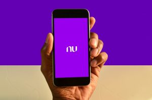 a imagem mostra uma mão segurando um smartphone e na sua tela há o logo do nubank há roxo ao fundo