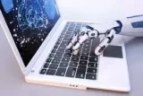 Imagem mostra mão robótica mexendo em notebook.