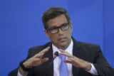Roberto Campos Neto, 53 anos, dá entrevista usando gestos com as mãos à frente de um fundo azul. Ele usa terno, óculos e gravata também azul.