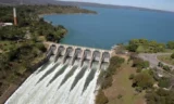 Imagem aérea mostra uma usina hidrelétrica com as comportas abertas em um dia ensolarado.