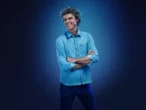 O tricampeão de tênis de Roland Garros, Gustavo Kuerten, posa para foto com sorriso no rosto, braços cruzados vestindo camisa e calça em tom azul sobre um fundo também azul