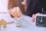 Pessoa coloca moeda dentro de cofre de porquinho posicionado entre miniatura de casa e um relógio