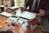 Profissional financeiro faz cálculos sobre o papel para cliente em frente, com miniatura de casa em cima da mesa