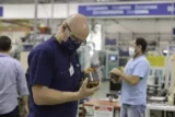 Imagem mostra funcionário no chão de fábrica de máscara inspecionando peça.
