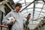 Jovem vibra vendo tela do celular enquanto segura bola de futebol. Ele veste moletom e gorro.