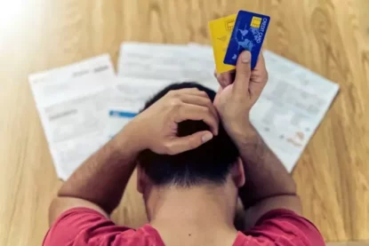 Rapaz leva as mãos à cabeça diante de boletos e dos cartões de banco que segura na mão