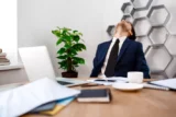 Jovem executivo de terno joga cabeça para trás em sinal de insatisfação diante de mesa de escritório.