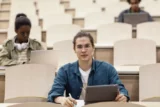 Estudante universitário em sala de aula, na frente de um notebook, pousa para a foto enquanto outros colegas estudam atrás.