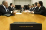 Placa em primeiro plano com a inscrição Copom e, desfocados no fundo, executivos engravatados sentados ao redor da mesa de reunião.