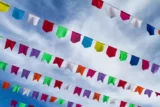 Bandeiras coloridas em corda pendurada na rua com céu de nuvens ralas ao fundo.