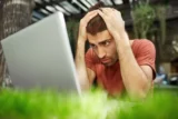 Jovem chateado e desapontado olhando triste para a tela do laptop sentado ao ar livre.