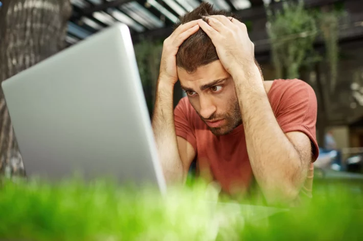 Jovem chateado e desapontado olhando triste para a tela do laptop sentado ao ar livre.