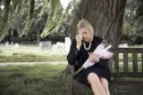 Mulher enxuga lágrimas sentada em banco de cemitério com buquê de flores na mão