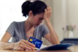 Mulher sentada à mesa, com uma mão na cabeça; e na outra mão, segurando um cartão bancário. Sobre mesa, há papéis de dívida.