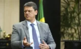 Homem de meia idade de terno gesticula durante discurso com bandeira do Brasil ao fundo.