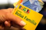 Imagem mostra detalhe de cartão do Bolsa Família e notas de reais na mão.