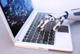 Imagem mostra mão mecânica de robô usando notebook