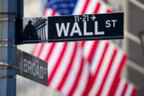 Placa de Wall Street, em Nova York, com bandeiras dos Estados Unidos ao fundo.