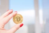 Imagem mostra mão segurando uma moeda de ouro representando o bitcoin.