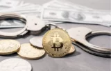 Imagem mostra brilhante moeda com símbolo do bitcoin destacada no centro em uma superfície com outras moedas e notas de dólar.