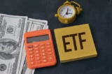 Imagem mostra notas de dólar dispostas ao lado de calculadora, relógio tipo despertador e uma placa escrito ETF.