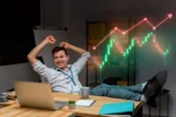 Imagem mostra jovem investidor relaxado com os pés sobre a mesa e sorrindo em frente a notebook. No fundo, gráficos que denotam ganhos com ativos do mercaddo financeiro. (Imagem: Freepik)