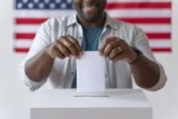 Cidadão sorridente coloca cédula na urna com bandeira dos Estados Unidos ao fundo.