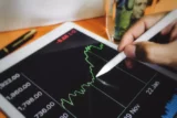Imagem mostra mão segurando uma caneta eletrônica sobre a tela de um tablet com um gráfico de linhas.