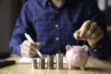 Imagem mostra rapaz colocando moeda dentro de cofre no formato de porquinho, com pilhas de outras moedas ao lado