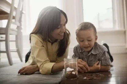 Imagem mostra mãe e filho deitados no chão contando moedas com um pote de vidro com várias delas.