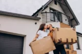 Imagem mostra jovem casal se beijando, com a fachada de casa ao fundo, enquanto seguram caixas de mudança.