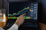 Imagem mostra mão apontando detalhe em um gráfico de ativo financeiro na tela de computador.