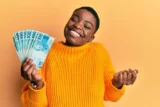 Imagem mostra jovem mulher de suéter sorrindo com notas de 100 reais na mão.