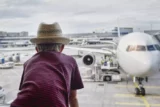 Imagem mostra garoto de chapéu em frente à janela vendo avião estacionado no pátio do aeroporto.