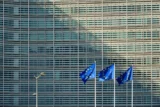 Imagem mostra bandeiras da União Europeia tremulando com prédio comercial ao fundo.