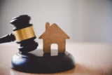 Imagem mostra martelo batendo sobre mesa com uma miniatura de casa ao lado.