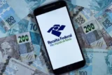 Imagem mostra celular na tela do aplicativo da receita Federal sobre superfície cheio de notas de 100 e 200 reais.