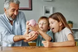 Imagem mostra criança esvaziando porquinho de moedas ao lado da família.