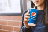 Imagem mostra jovem mulher bebendo refrigerante de canudinho, em copo da Pepsi.