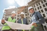 Imagem mostra três trabalhadores concentrados em um desenho de projeto com construção de prédio ao fundo.