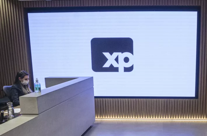 Imagem mostra recepção de escritório da XP, formado por um balcão com uma atendente e a logo da empresa em destaque na parede.