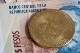 Imagem mostra moeda representativa do bitcoin sobre nota de pesos argentinos.