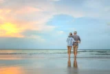 Imagem mostra casal de aposentados em caminhada na praia, abraçados.
