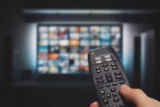 Imagem mostra controle na mão com tela de TV ao fundo.