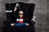Garoto de óculos escuros e jaqueta de couro está sentado em uma poltrona grande com um monte de dólares em dinheiro, jogando as notas para o alto.